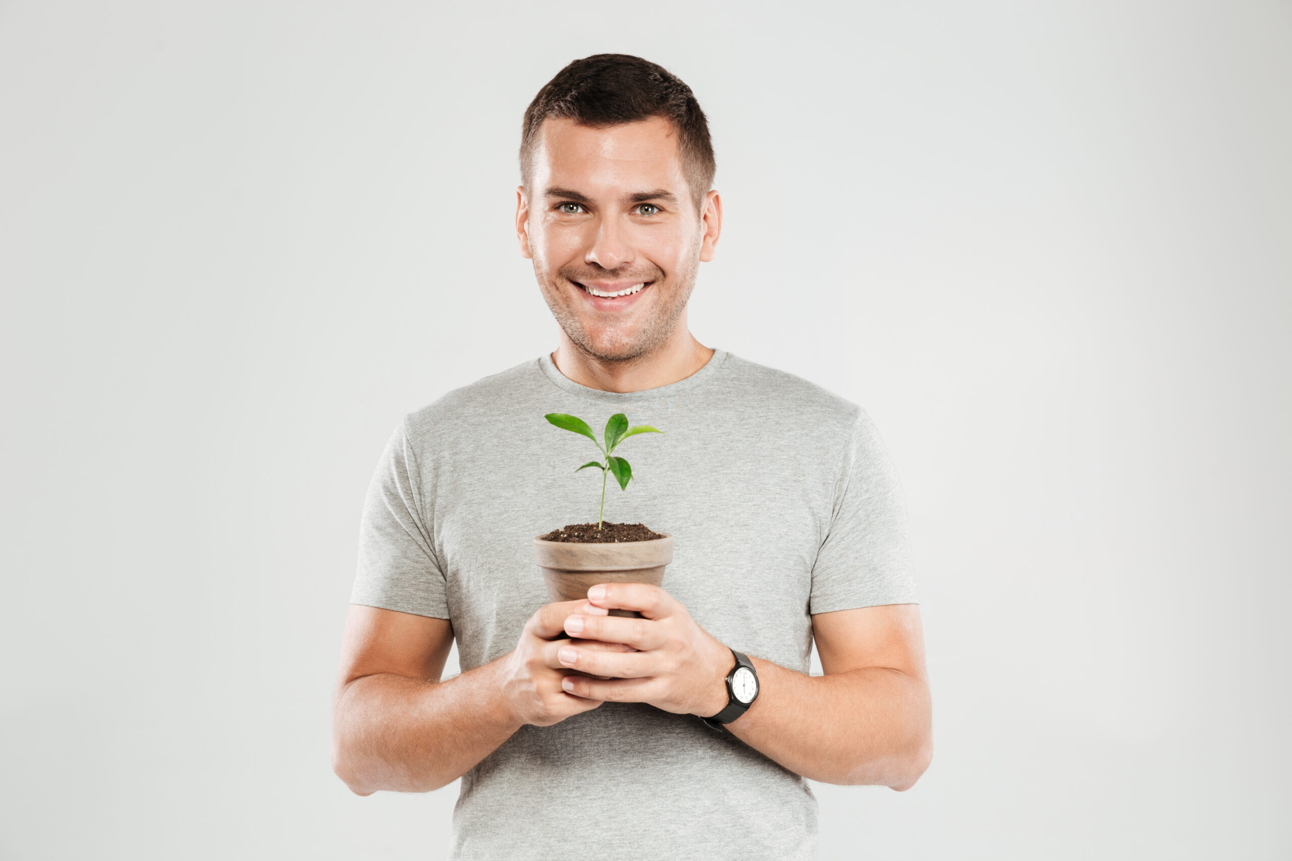 Photo of young smiling man dressed in grey t-shirt isolated over grey wall background. Looking camera while holding plant.