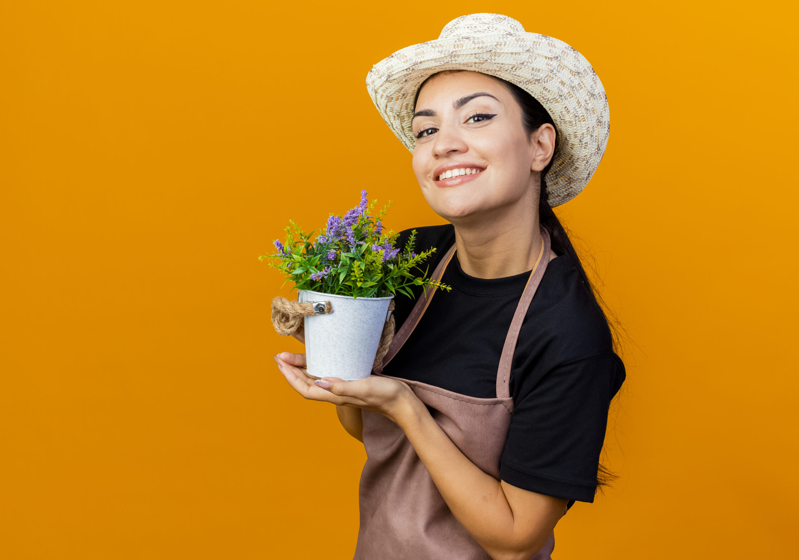 young beautiful woman gardener in apron and hat holding potted plant looking at camera smiling standing over orange background